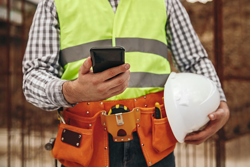 Male construction worker from the neck down wearing yellow safety vest, an orange tool belt, holding a white hard hat and holding a black smartphone.