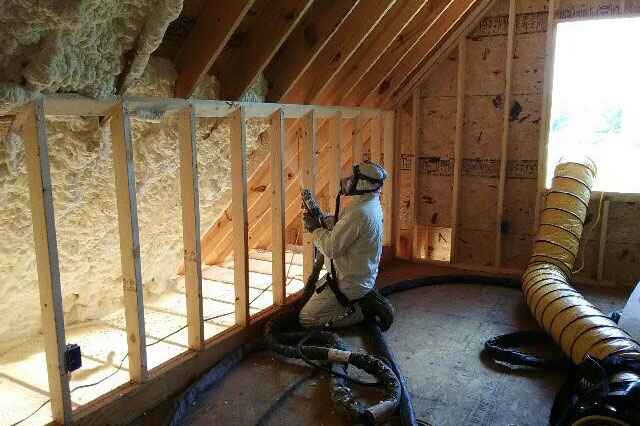 Man in white full body suit installing spray foam insulation in an attic.