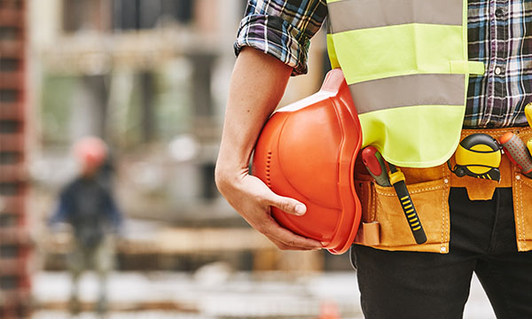 Construction worker. Cropped photo of male professional builder in working uniform with construction tool belt, yellow safety vest, holding a safety helmet while standing outdoor of construction site.