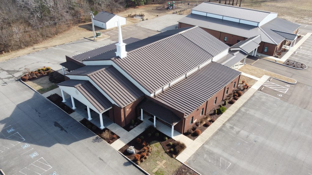 An aerial view of the newly-built Grace Baptist Church. The roof shows a dark brown color and the church is surrounded by a large parking area. A storage building is seen on the upper left side.