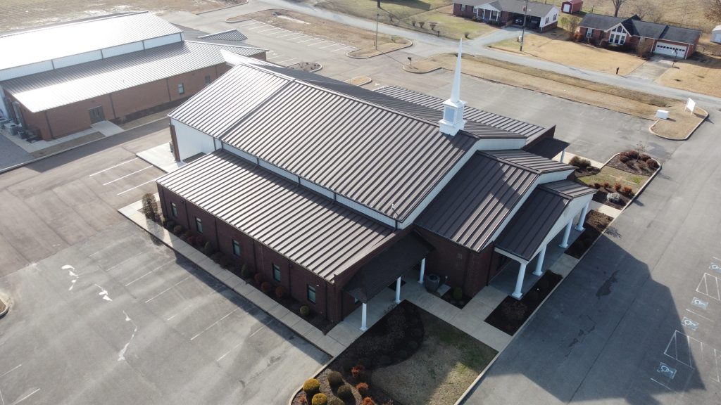 An drone shot of the newly built Grace Baptist Church with a dark brown metal roof. A wide parking area can be seen around the church and a wooded area can be seen on the upper right side of the church. Residential homes can be seen on the upper right side.