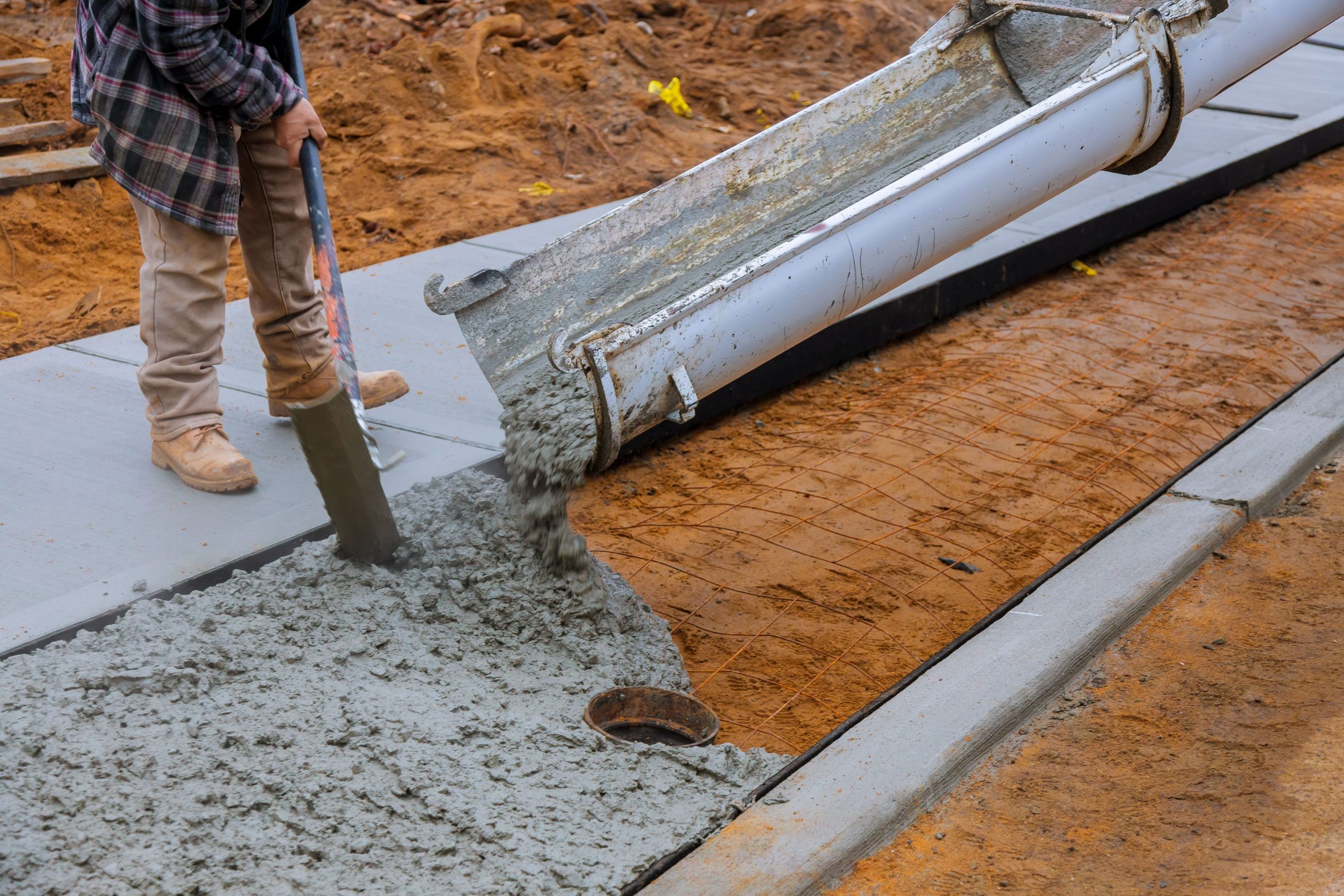 A construction worker wearing checkered jacket and caterpillar boots guiding the flow of liquid concrete on reinforcing bars.