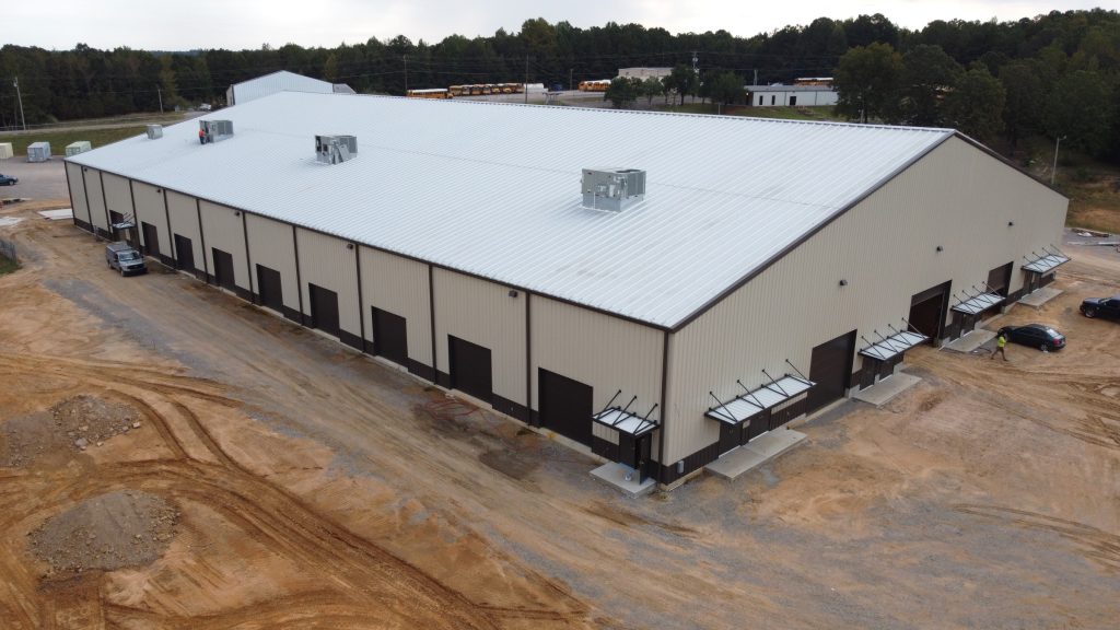 A drone shot of the exterior side area of the newly built McNairy County Agricultural Event Center. A black car and black truck can be seen parked on the back area. A heavily-wooded area and some buildings can be seen on the far distant area.
